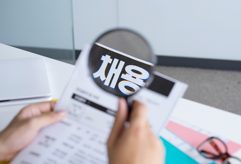 한 사람이 '채용'이라고 쓰여진 종이를 보고 있는데, 채용이라는 글자에 돋보기를 대고 있어 글자가 크게 보인다.