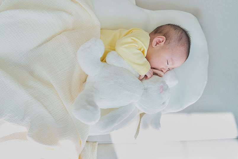 하얀 침구류 위에서 인형을 안고 '수면'을 취하고 있는 아기의 모습이다.